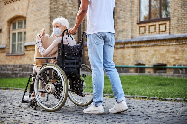 Dame âgée handicapée tenant un sac en papier avec des achats et faisant demi-tour pendant qu'une jeune pousse son fauteuil roulant