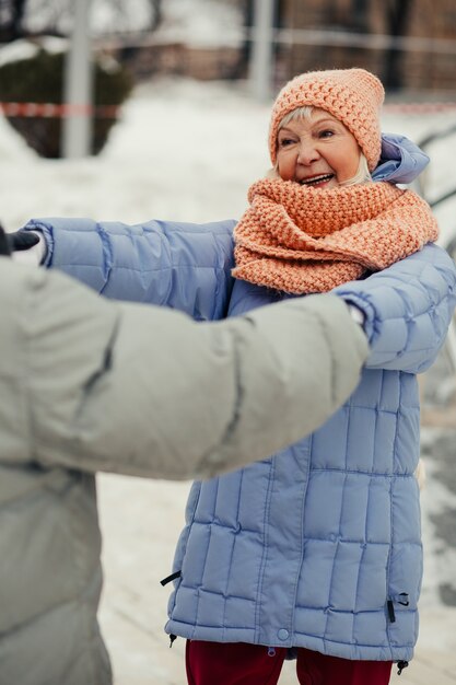 Photo dame âgée en manteau d'hiver souriant à son mari tout en lui tenant les mains et en dansant avec lui dans la rue