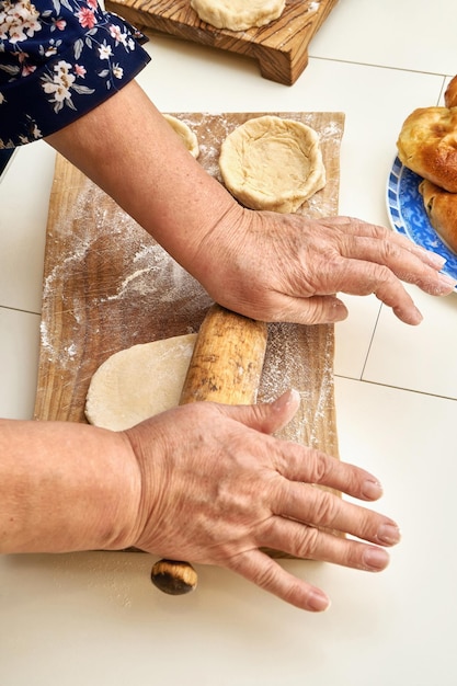 Dame âgée roule un morceau de pâte crue avec un rouleau à pâtisserie faisant cuire des tartes maison à la table blanche vue rapprochée