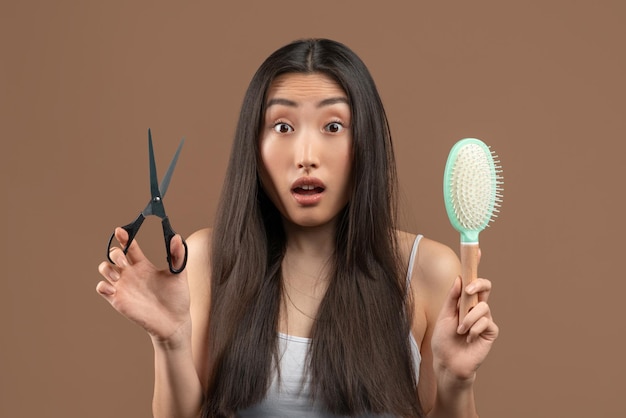 Dame asiatique choquée aux cheveux longs tenant un peigne et des ciseaux en métal pensant à un traitement professionnel