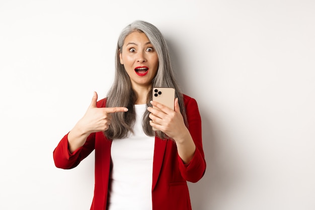 Dame asiatique étonnée en blazer rouge pointant le doigt sur un smartphone, regardant impressionnée par la caméra, montrant quelque chose en ligne, fond blanc