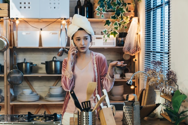 une dame asiatique malheureuse en pyjama avec une serviette sur la tête porte une expression confuse tout en parlant au téléphone avec un geste de paume vers le haut dans la cuisine à la maison