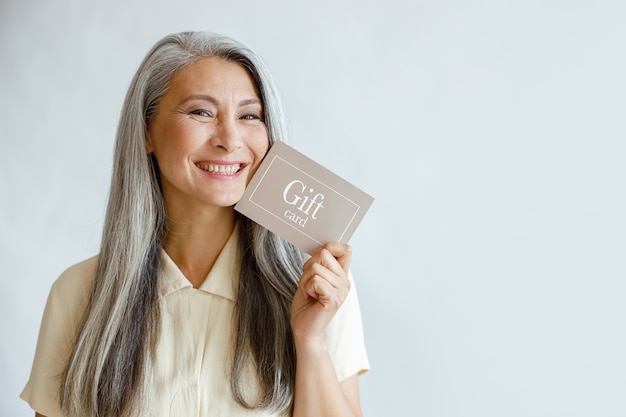 Une dame asiatique mature assez souriante tient une carte-cadeau posant sur fond gris clair en studio, espace pour le texte. Certificat d'achat