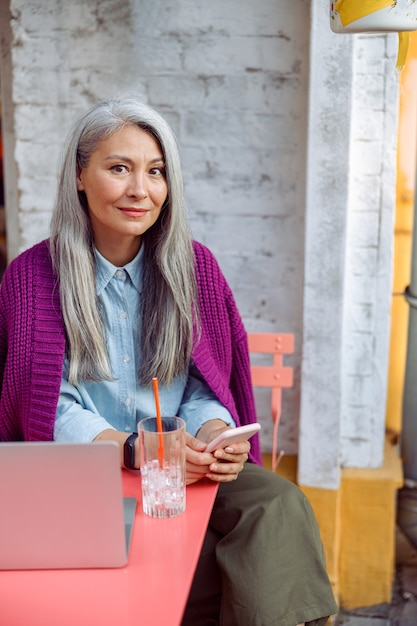 Une dame asiatique mature et positive aux cheveux raides tient un téléphone portable à table avec un ordinateur portable