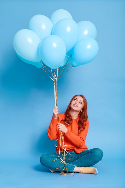 Dame au gingembre portant un pull orange tenant des ballons d'hélium bleu et regardant avec le sourire, femme assise sur le sol et célébrant son anniversaire, posant isolé sur un mur de couleur.