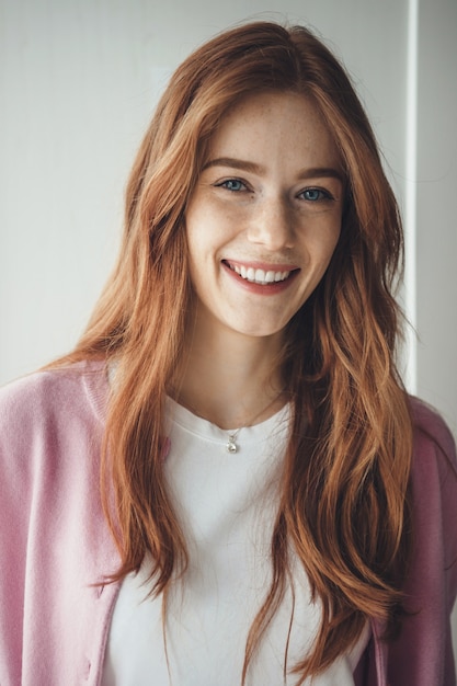 Dame au gingembre avec des taches de rousseur souriant à pleines dents à la caméra portant un t-shirt blanc et un pull rose