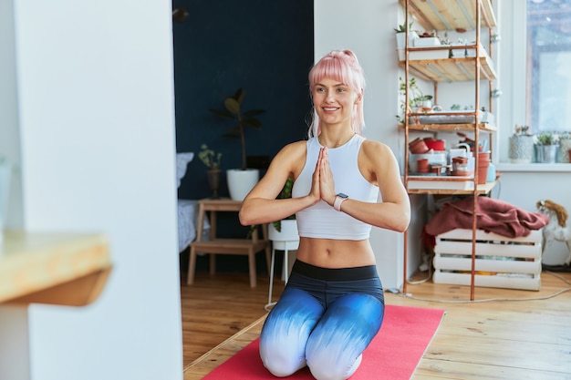 Une dame aux cheveux roses souriante tient la main dans namaste mudra assise sur les genoux à la maison