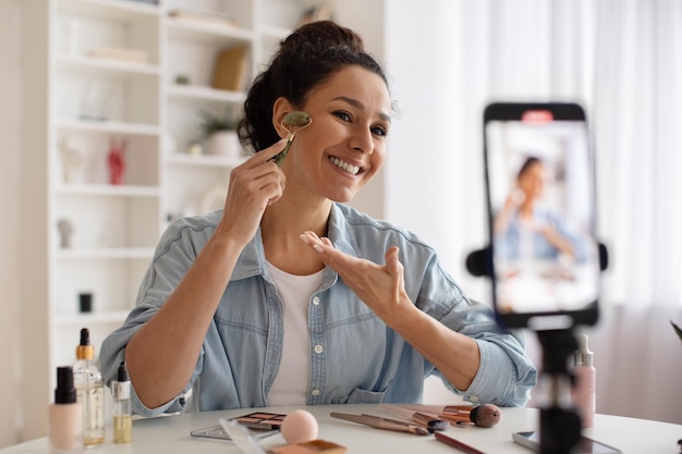 Dame blogueuse faisant un massage facial à l'aide d'un rouleau faisant une vidéo à l'intérieur