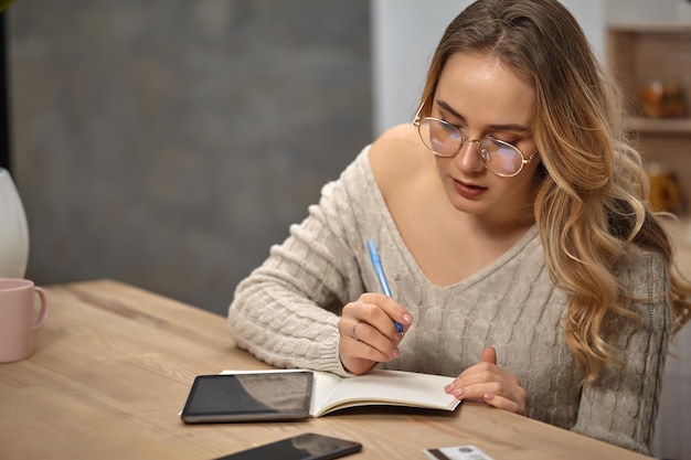 Dame blonde blogueuse en chandail tricoté beige à lunettes Assis dans la cuisine à une table en bois avec une tasse rose dessus Écrit quelque chose dans un ordinateur portable Tablette smartphone et carte en plastique à proximité Gros plan