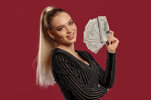 Dame blonde en robe noire en strass montrant un fan de billets de cent dollars souriant posant sur le côté...