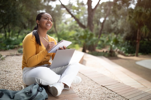 Dame brésilienne joyeuse assistant à un webinaire à l'extérieur assis dans un parc à l'aide d'un ordinateur portable et prenant des notes