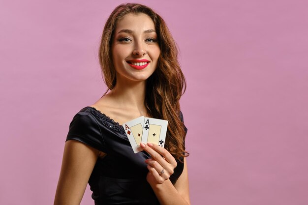 Dame brune avec boucle d'oreille dans le nez, en robe noire. Montrant deux cartes à jouer, souriant, posant sur fond de studio rose. Poker, casino. Fermer