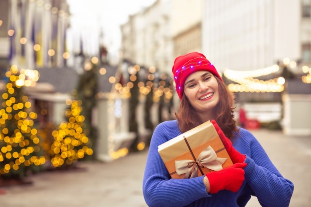Une dame brune joyeuse porte un chapeau rouge et un pull bleu tenant une boîte-cadeau sur la foire de Noël décorée de guirlandes. Espace pour le texte