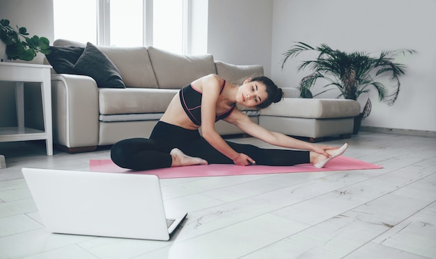 Dame brune en tenue de sport qui s'étend sur le sol en regardant l'ordinateur portable et en faisant des exercices de yoga