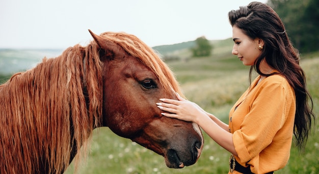 Dame brune touchant son cheval brun tout en posant dans un champ près de la forêt