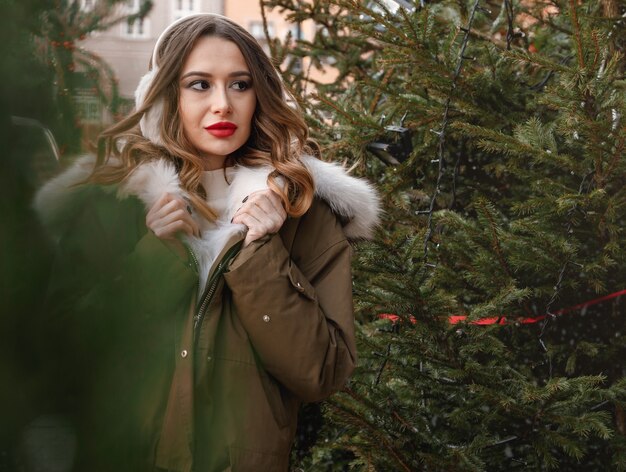 Dame caucasienne dans un pull en tricot et des écouteurs moelleux. Portrait en plein air de la belle jeune fille souriante heureuse porte un manteau de fourrure près de l'arbre de Noël décoré. Pologne