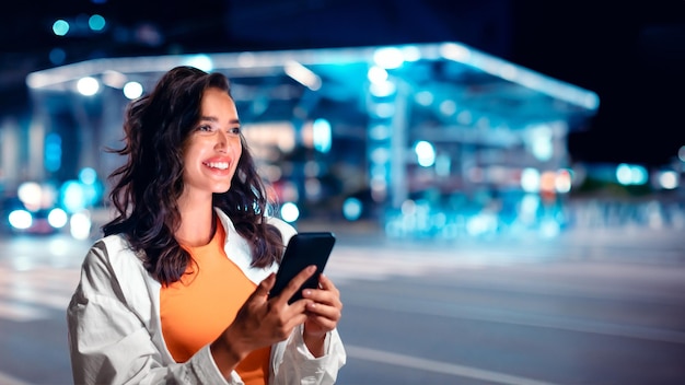 Dame caucasienne excitée utilisant un smartphone tout en marchant dans la rue nocturne de la ville pleine de néons