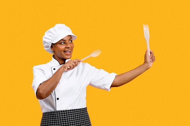 Dame de chef noire ludique avec des spatules dans les mains posant sur fond jaune