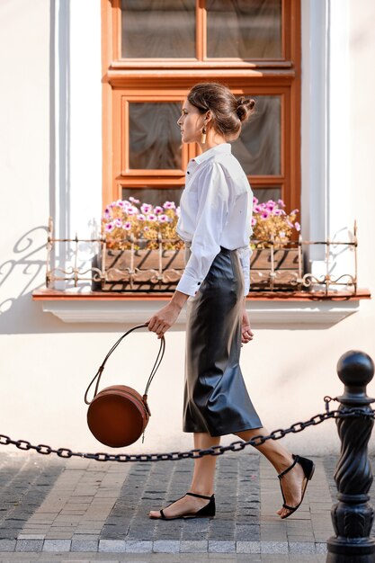 Dame élégante En Jupe En Cuir Et Chemise Blanche Marchant Dans La Rue Avec Un Sac à Main En Cuir