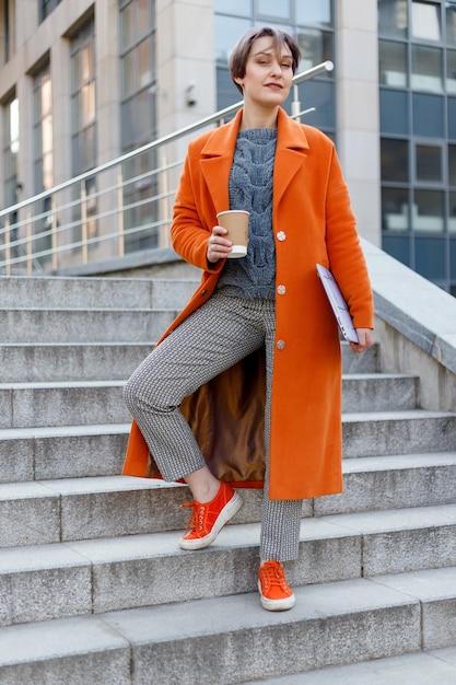 Dame élégante en manteau orange vif avec son ordinateur portable et sa tasse de café