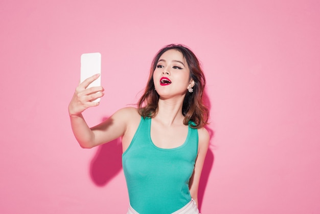 Dame d'été. Belle fille asiatique avec maquillage professionnel et coiffure élégante prenant une photo de selfie sur fond rose.