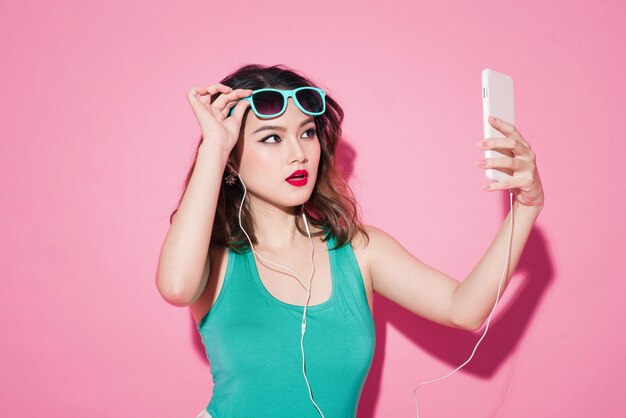Dame d'été. Belle fille asiatique avec maquillage professionnel et coiffure élégante prenant une photo de selfie sur fond rose.