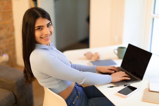 Dame étudiante espagnole joyeuse assise au bureau et utilisant un ordinateur portable avec écran blanc souriant à