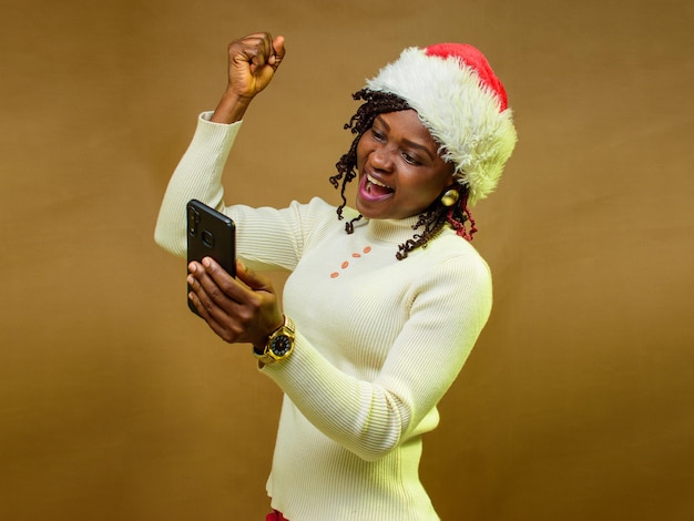 Une dame ou une femme africaine regardant joyeusement le téléphone intelligent dans sa main et ayant également une casquette de Noël sur la tête