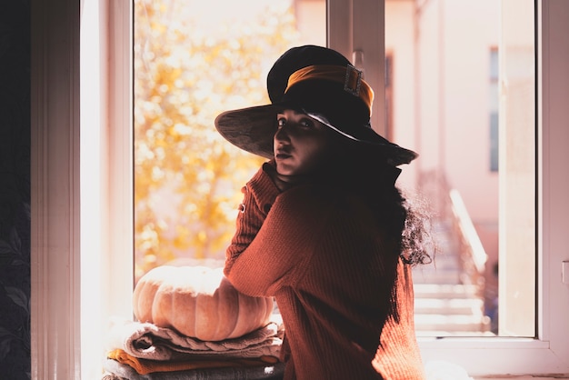 Photo dame d'halloween portant une coupe de sorcière une femme en chapeau de sorcière et citrouille d'halloween près des fenêtres portrai...