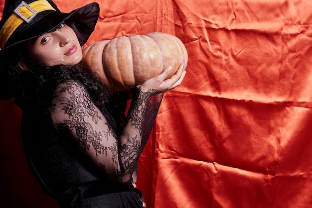 Photo dame d'halloween portant une tasse de sorcière et tenant une grosse citrouille d'halloween une femme au chapeau de sorcière et sanctifier...