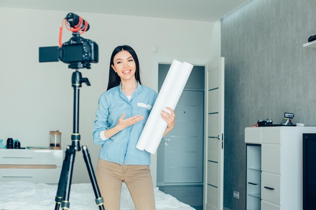 Dame joyeuse faisant des gestes et souriant devant une caméra en se tenant debout avec un rouleau de papier et en parlant devant une caméra