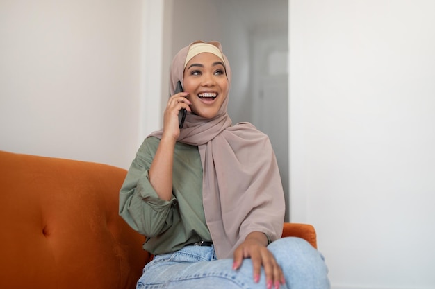 Dame joyeuse en hijab parlant au téléphone portable assise à la maison