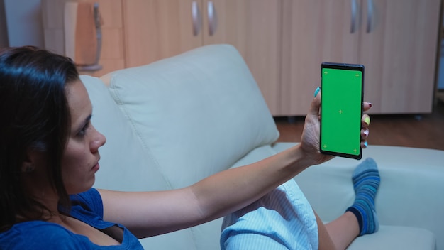 Dame lisant sur un écran de smartphone avec écran vert allongé sur un canapé dans le salon. Femme tenant un téléphone portable avec un modèle de maquette chroma key écran de téléphone portable isolé à l'aide d'internet techology