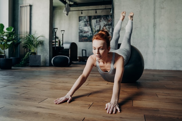 Dame mince athlétique aux cheveux roux effectuant des exercices d'étirement sur un grand fitball argenté dans une ambiance de fond de studio de fitness, se concentrant sur l'exercice pour la force et l'équilibre