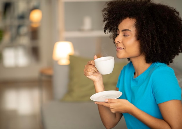 Dame noire bouclée millénaire positive tenir une tasse de thé profiter de l'arôme bon temps libre savoureux sur le canapé