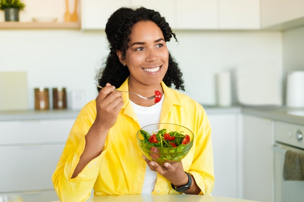 Dame noire positive mangeant une salade de légumes frais pour le dîner assis dans un intérieur de cuisine clair à la maison espace libre