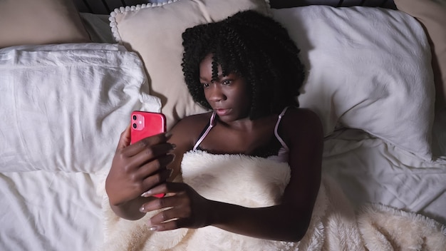Une dame noire sérieuse regarde un smartphone rouge moderne et des types allongés sur un lit queen-size dans la chambre la nuit