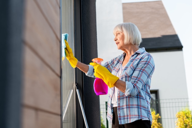 Dame occupée. Vieille belle dame portant des gants jaune vif se sentant extrêmement occupé à laver les vitres