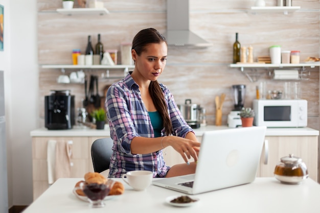 Dame pointant sur l'écran de l'ordinateur portable pendant la matinée dans la cuisine avec petit-déjeuner à côté d'elle et tasse de thé vert