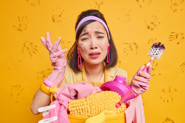 dame porte des gants de protection en caoutchouc bandeau tablier tient une brosse de toilette sale se tient près d'un panier à linge avec des pinces à linge et du détergent isolé sur jaune