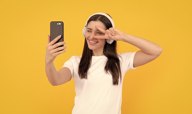 Dame positive en chemise blanche dans les écouteurs faisant selfie au téléphone avec selfie geste de paix