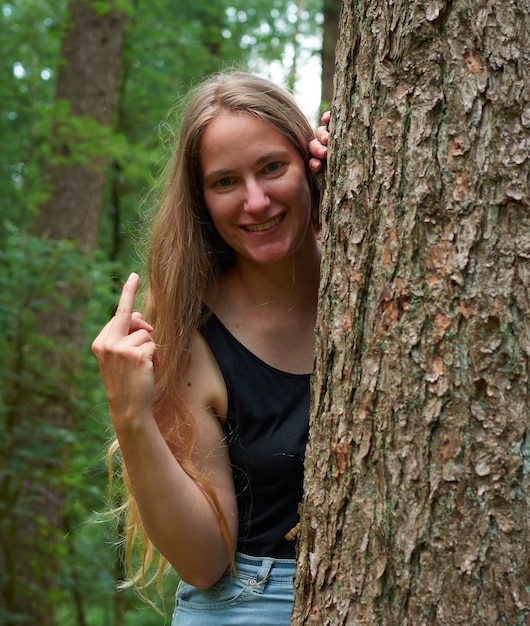 Une dame de race blanche blonde aux cheveux longs à la recherche de derrière un tronc d'arbre montrant un doigt du milieu