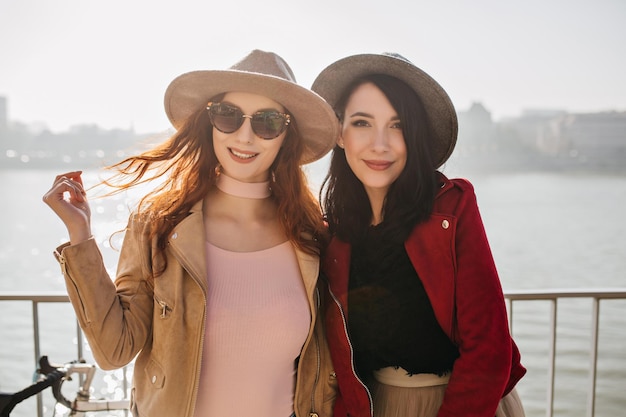 Dame romantique en veste beige joue avec les cheveux longs en posant avec un ami. Deux filles blanches en chapeaux profitant d'une vue sur la mer.