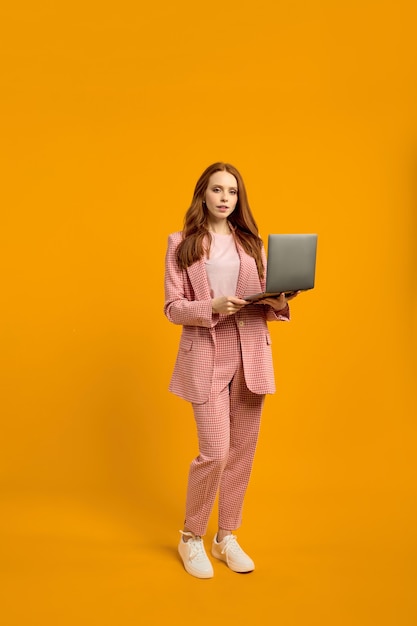 Dame rousse au gingembre confiant marchant avec un ordinateur portable isolé sur un dos de studio orange jaune vif...