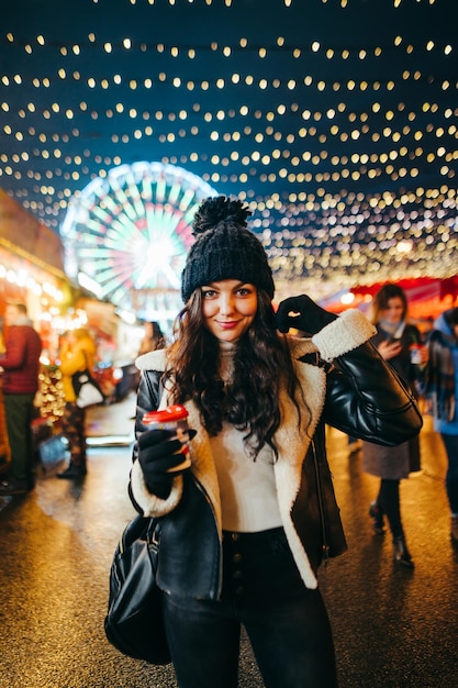 dame se promène le soir dans la rue décorée pour Noël avec une tasse de boisson chaude à la main