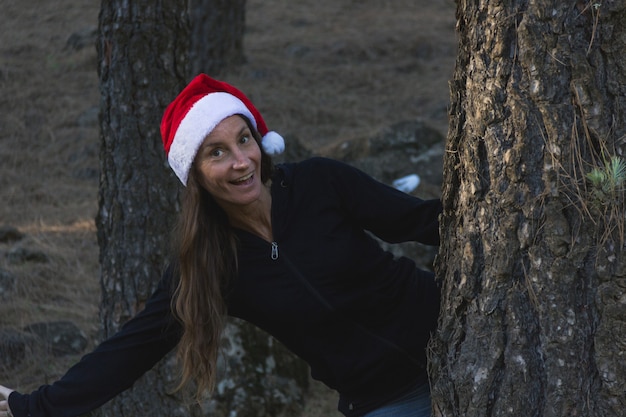 Dame souriante avec chapeau rouge de Noël tenant sur un tronc de pin dans la forêt Heureuse femme d'âge moyen