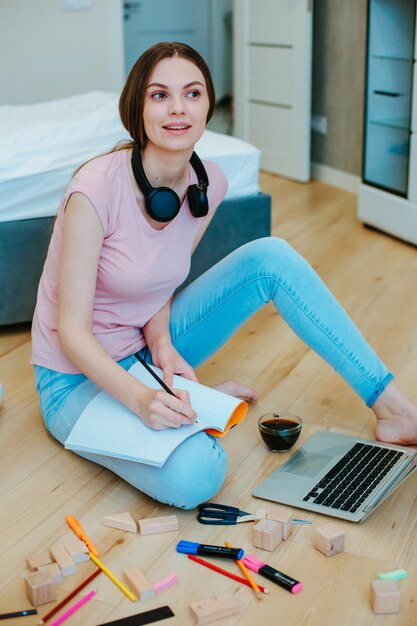 Dame souriante en tenue décontractée assise sur le sol avec un ordinateur portable et regardant ailleurs tout en prenant des notes