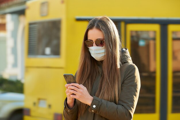 Une dame utilisant un smartphone porte un masque médical pour éviter la propagation du coronavirus dans une rue de la ville. Une fille à lunettes de soleil avec un masque chirurgical sur le visage contre COVID-19.