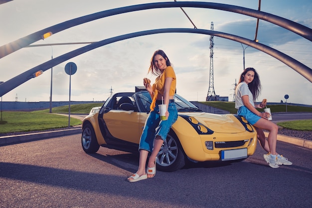 Les dames sourient en tenant des hamburgers et des boissons dans des gobelets en papier tout en étant assises sur le capot de la voiture jaune