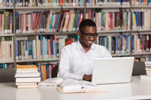 Dans la bibliothèque beau étudiant africain avec ordinateur portable et livres travaillant dans une bibliothèque universitaire de lycée faible profondeur de champ